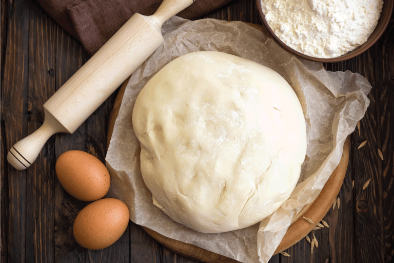 fresh dough left on a table to rise with eggs, rolling pin, and flour. How Long To Defrost Dough - By Type Of Dough