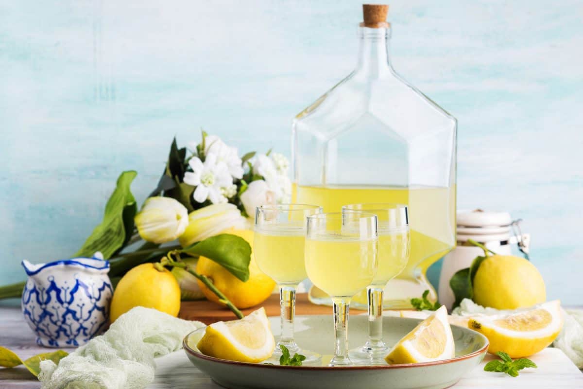 Three small glasses and a huge jar of lemoncello with lemons and flowers on top of a table